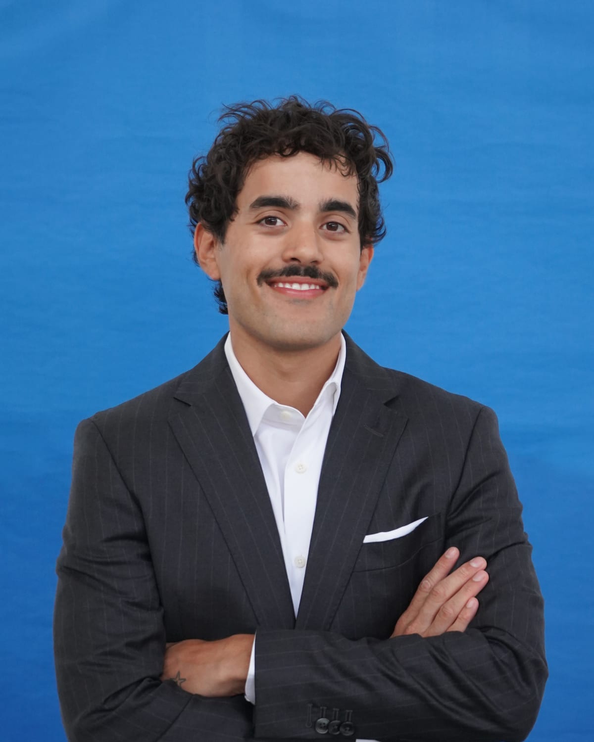 Retrato de estúdio do Lucas Viegas, de fato escuro e camisa branca, num fundo azul.