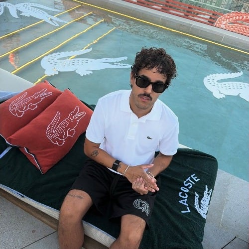 O Lucas sentado à beira de uma piscina, com toalhas da Lacoste.